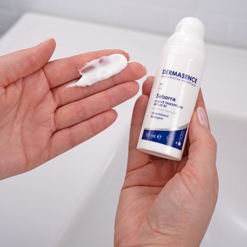 Hand toont een pompflacon Dermasence Seborra Light day cream SPF 30; op de andere hand ligt een streep witte gelcrème, bij een wastafel.
