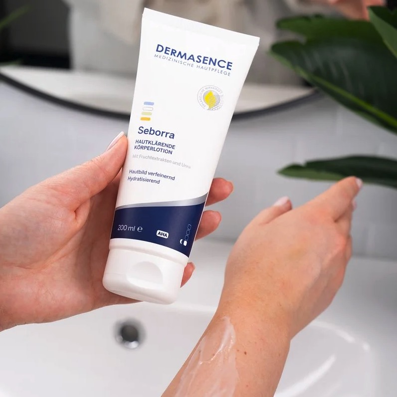 Hand die een tube DERMASENCE Seborra huidklarende bodylotion vasthoudt en de lotion op de onderarm aanbrengt bij een wastafel.