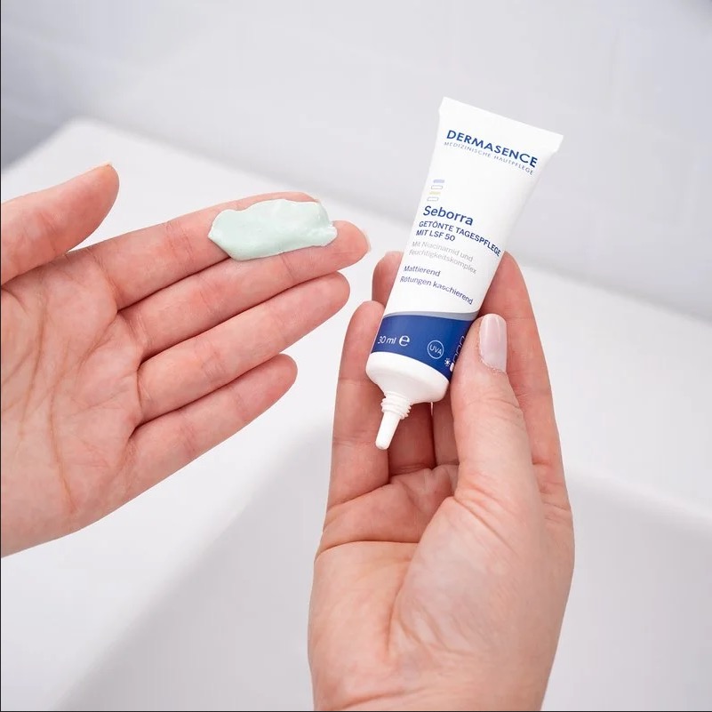 Een hand met een beetje lichtgroene crème uit een tube Dermasence Seborra Tinted Day Cream SPF