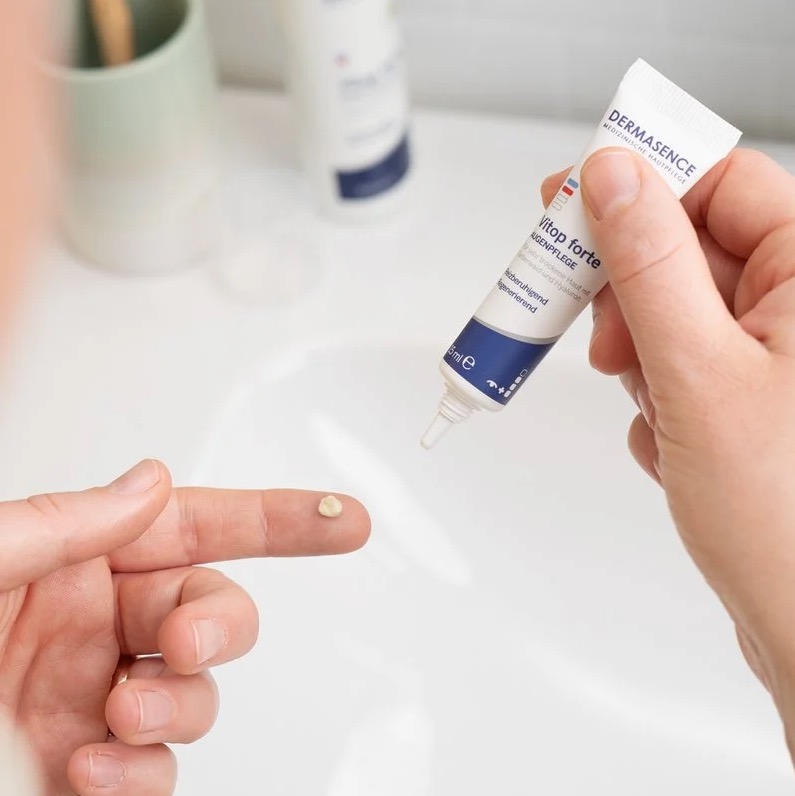 Een persoon brengt een kleine hoeveelheid beige oogcrème aan op een vingertop met een tube DERMASENCE Vitop Forte oogcreme erboven gehouden, in een lichte badkameromgeving.