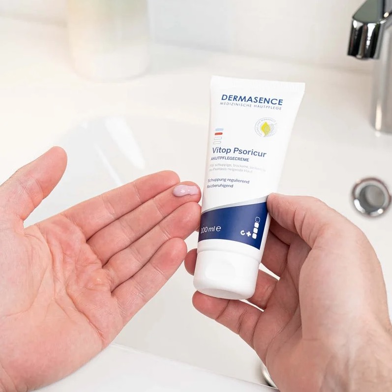 Een persoon houdt een tube DERMASENCE Vitop Psoricur creme vast en brengt een kleine hoeveelheid witte crème aan op de vingertop, boven een wastafel.