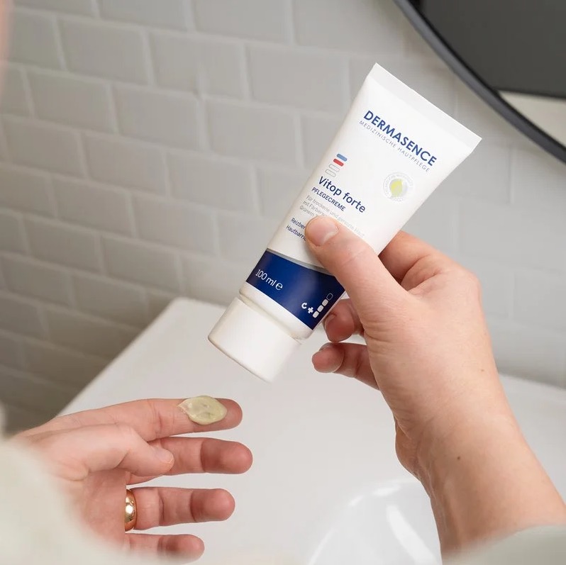 Een persoon knijpt een beetje beige-groene crème uit een tube DERMASENCE Vitop Forte creme op een vingertop, boven een wastafel met witte tegels op de achtergrond.