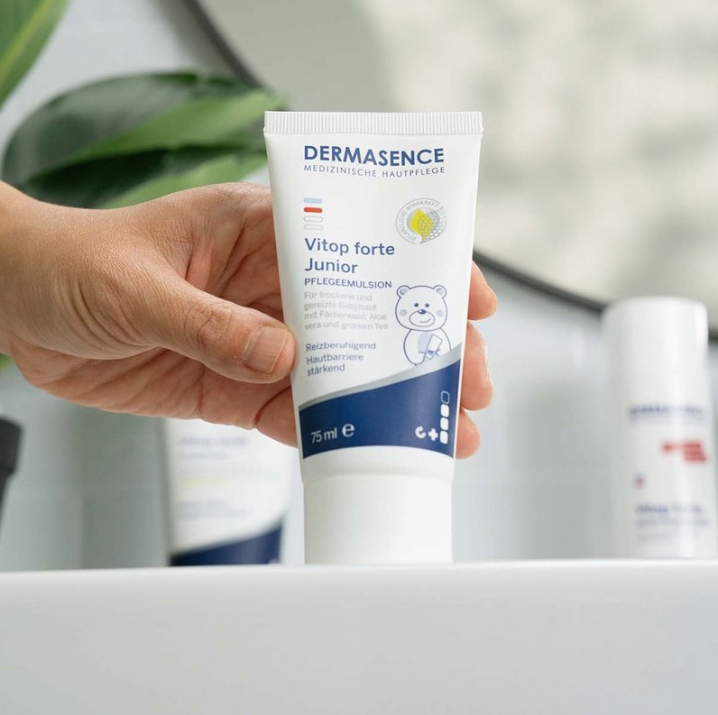 Een hand houdt een tube DERMASENCE Vitop Forte Junior creme vast. De tube toont een illustratie van een beertje en is bedoeld voor droge, gevoelige baby- en kinderhuid.