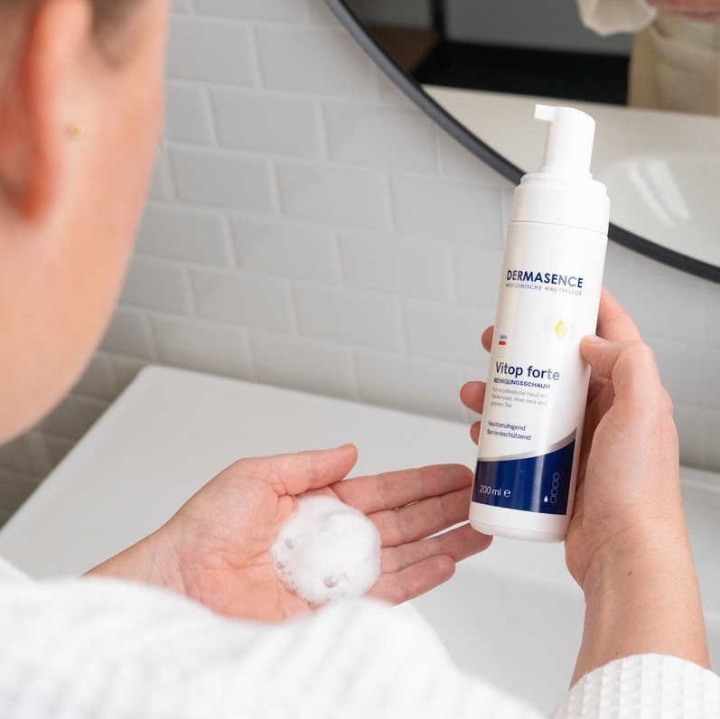 Een persoon pompt een hoeveelheid wit reinigingsschuim in de hand uit een fles DERMASENCE Vitop Forte Cleansing Foam, tegen een achtergrond van witte badkamertegels.