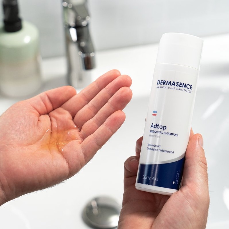 Een persoon houdt een fles DERMASENCE Adtop Medizinal Shampoo vast terwijl een kleine hoeveelheid goudkleurige shampoo in de andere hand ligt, boven een witte wastafel.