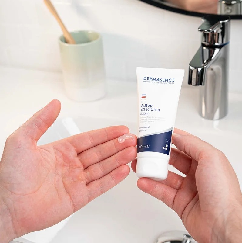Een persoon houdt een tube DERMASENCE Adtop 40% Urea Oleogel vast en heeft een kleine hoeveelheid transparante gel op de handpalm, boven een witte wastafel.