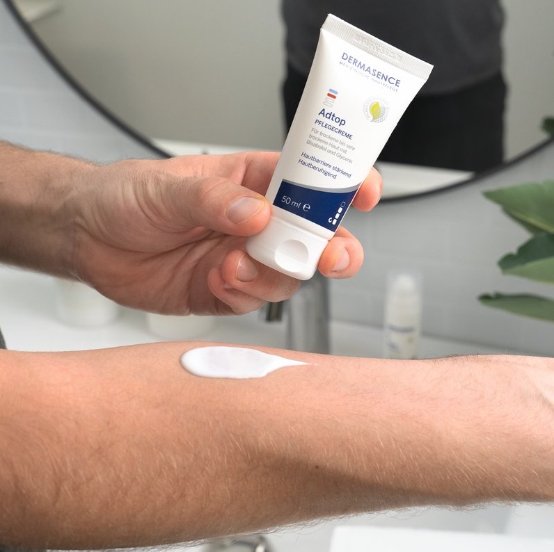 Een persoon brengt een royale streep witte crème aan op de onderarm vanuit een tube DERMASENCE Adtop carecream, in een badkameromgeving.