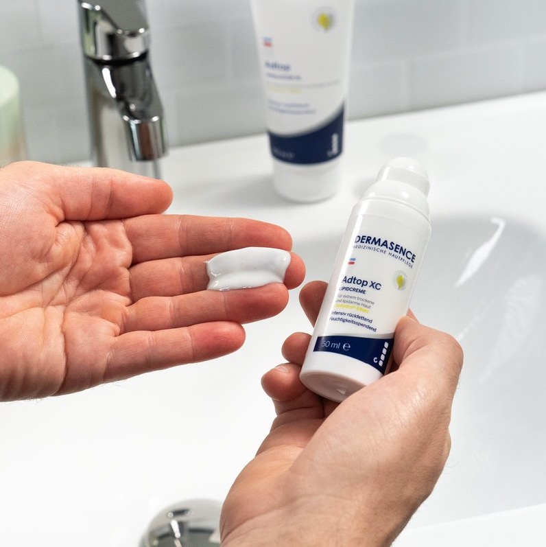 Een persoon houdt een pompflacon DERMASENCE Adtop XC Lipidcreme vast en heeft een royale hoeveelheid witte, rijke crème op de handpalm, boven een wastafel met badkamertegels.