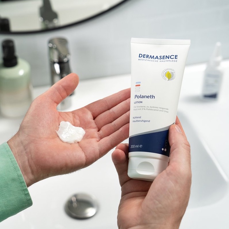 Een persoon houdt een tube DERMASENCE Polaneth Lotion vast terwijl een royale hoeveelheid witte lotion in de andere hand ligt, boven een wastafel in een badkamer.
