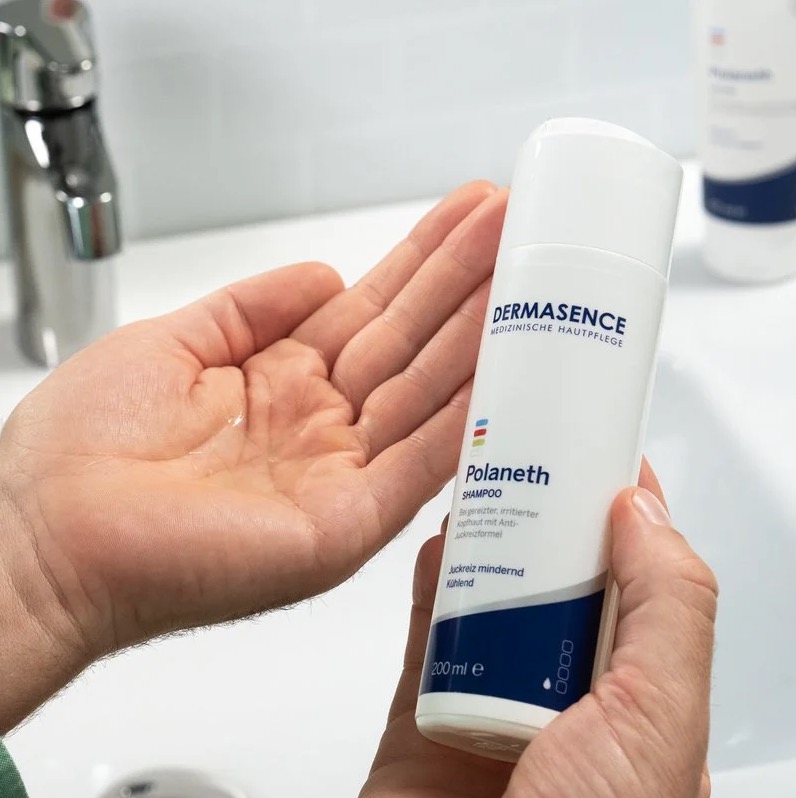 Een persoon houdt een fles DERMASENCE Polaneth Shampoo vast terwijl een kleine hoeveelheid transparante shampoo in de hand ligt, boven een wastafel met witte tegels.