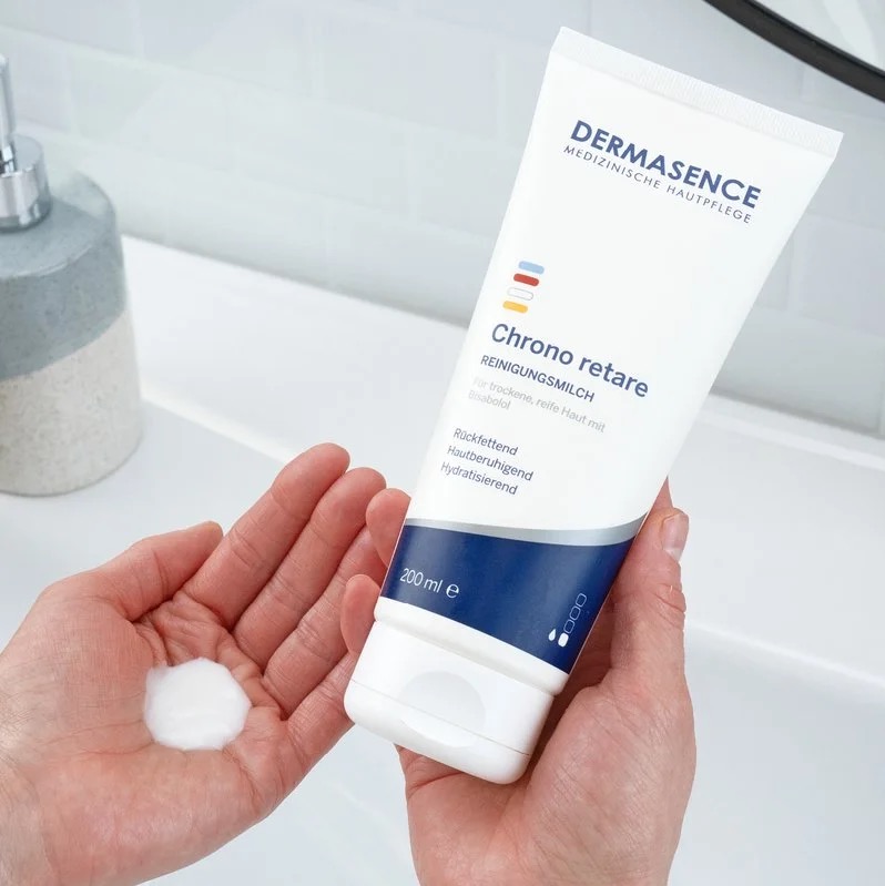 Handen tonen een tube Dermasence Chrono retare Reinigingsmelk; in de andere hand ligt een dot witte reinigingsmelk, bij een wastafel in de badkamer.