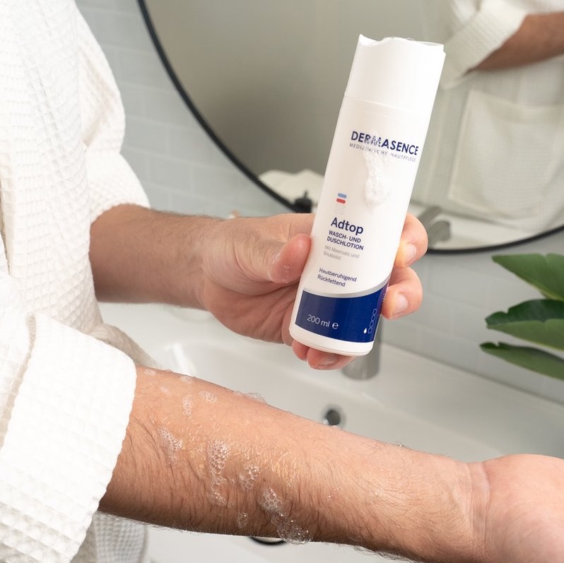 Een persoon wrijft schuimende DERMASENCE Adtop Waschlotion uit over de onderarm terwijl de fles in de andere hand wordt vastgehouden, in een badkamer met witte tegels en wastafel op de achtergrond.